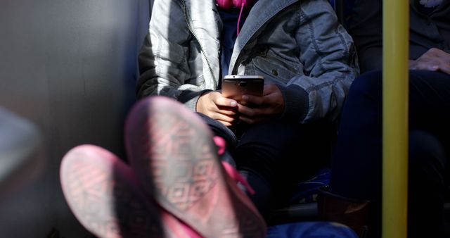 Person wearing casual clothing sits on public transport while using smartphone. Pink shoes casually propped up. Can be used for concepts of daily commute, modern technology, relaxation during travel, and everyday travel experiences.