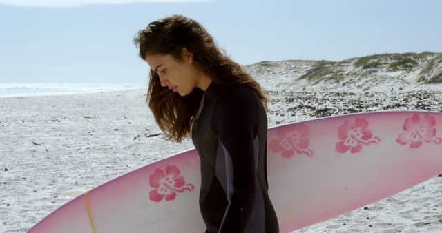 Female surfer in a wetsuit walking along a sandy beach while holding a pink surfboard with floral design. Ideal for use in websites and materials related to surfing, outdoor sports, beach activities, seaside tourism, and fitness.