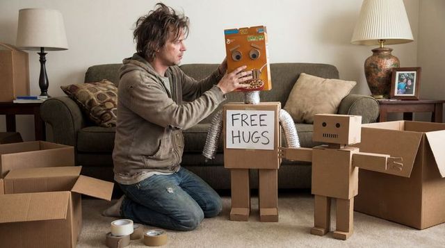 Kneeling man placing cereal-box head on cardboard robot holding free hugs sign in living room