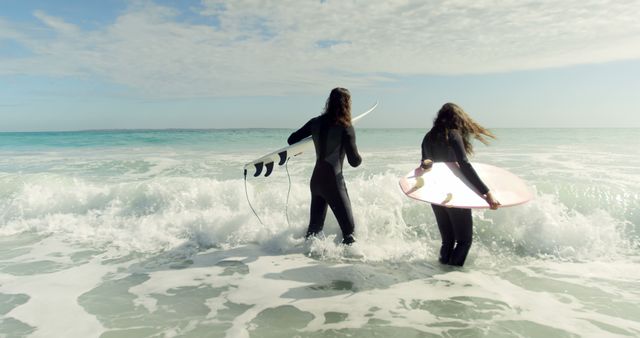 Two female surfers are entering the ocean, carrying their surfboards, wearing wetsuits. This image can be used for promoting beach vacations, surf schools, water sports equipment, outdoor adventure lifestyle, or female empowerment in sports.