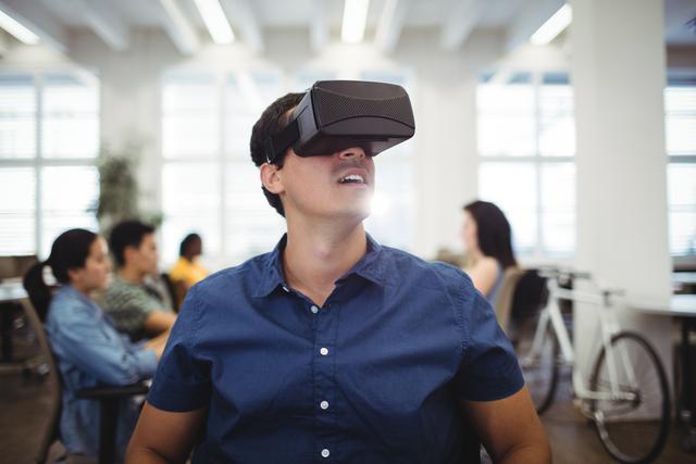Man using virtual reality headset in a modern office environment, surrounded by colleagues. Ideal for illustrating concepts related to digital innovation, tech-savvy workplaces, and immersive technologies in business settings.