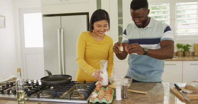 Happy multiracial couple standing in a modern kitchen preparing breakfast together. The man is cracking eggs into a bowl while the woman holds a jug of milk. Both are smiling and enjoying their time together. This image is perfect for depicting bonding moments, family life, cooking tutorials, and lifestyle or relationship articles.