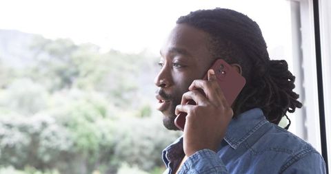 African American man speaking on smartphone by window wearing denim shirt and nose stud