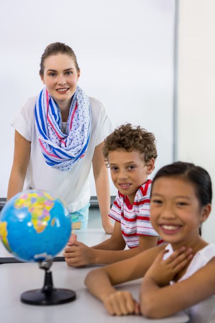 Teacher with two diverse students sitting at a table in a classroom. The teacher is standing behind the students, who are smiling and looking at the camera. A globe is on the table, indicating a geography lesson. Ideal for educational materials, school brochures, and websites promoting diversity and inclusion in education.