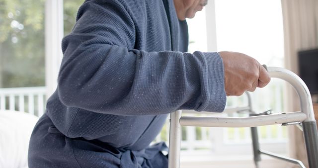 Elderly man using a walker for support indoors, focusing on maintaining balance and mobility. This image is useful for illustrating senior health care, mobility aids, rehabilitation, and independent living concepts. It is ideal for use in medical articles, healthcare brochures, elder care service advertisements, and wellness blogs.