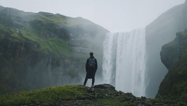 Male hiker standing at misty canyon edge facing towering waterfall, exploring remote wilderness