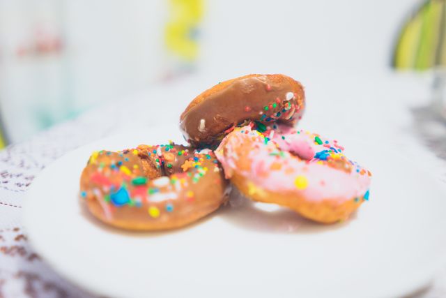 Three vibrant doughnuts with multicolored sprinkles are tastefully arranged on a white plate. Ideal for food blogs, bakeries, cafes, or promotional materials emphasizing treats and desserts. Also, perfect for social media posts focused on delicious sweets.