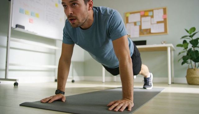 Man performing plank on mat in home office wearing smartwatch, blue tee, focused workout