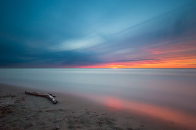 Driftwood resting on the beach against a backdrop of a colorful sunset. Sky features vibrant hues of blue, pink, and orange. Calm water and smoothing effect on waves due to long exposure make this scene tranquil. Perfect for use in nature-themed projects, relaxation visuals, or travel advertisements.