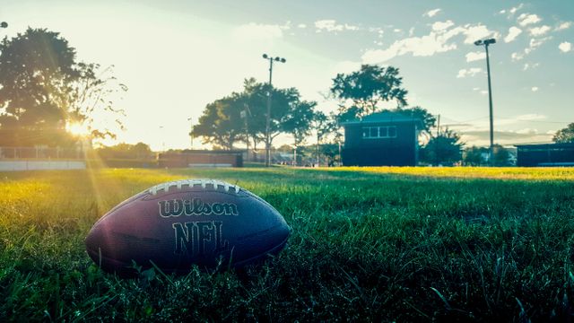 American football lying on a grassy field under the warm glow of the setting sun. This serene and picturesque scene captures the essence of outdoor sports and recreation. Ideal for use in articles, advertisements, and posters related to sports, relaxation, outdoor activities, and evening events.