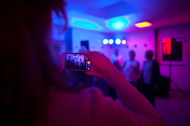 Person is capturing a group of friends using a smartphone during a party. Colorful lights add a vibrant and lively atmosphere to the scene. This can be used for themes such as night life, celebrations, social gatherings, or memories at a party.