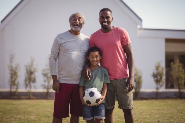 This image shows a happy multi-generation family standing in a garden with a football. The grandfather, father, and son are smiling and enjoying their time together. This photo is ideal for use in advertisements, family-oriented content, and articles about family bonding, outdoor activities, and sports.