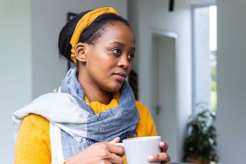 African american woman holding mug by window wearing yellow sweater, gray scarf