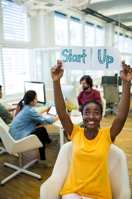 Young female entrepreneur holding a 'Start Up' sign, smiling in a modern office. Background shows a diverse team working on computers, suggesting a collaborative and innovative environment. Ideal for use in articles or advertisements about entrepreneurship, start-ups, business success, teamwork, and modern workspaces.