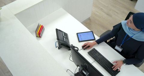 Female receptionist wearing hijab managing modern front desk with computer and tablet