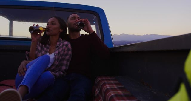 Couple sitting in back of pickup truck enjoying time together while drinking beverages and watching sunset. Perfect for themes of love, togetherness, nature outings, road trips, and relaxation.