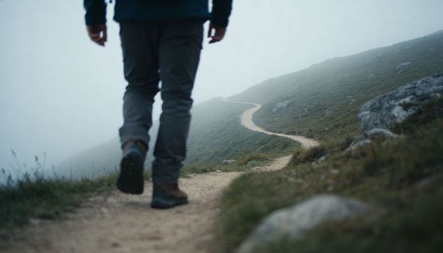 Solo hiker walking on winding misty hillside trail with rugged boots and rocky path