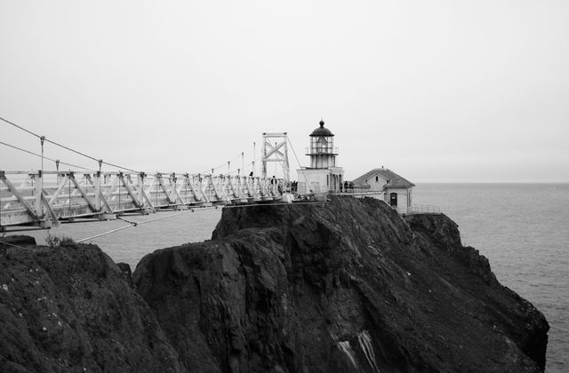 Features lighthouse perched on edge of rocky cliff with a suspended bridge in black and white setting. Ideal for themes related to maritime history, coastal landscapes, seafarers, and nostalgia. Suitable for use in postcards, wall art, educational materials, and travel blogs focusing on navigation and lighthouses.