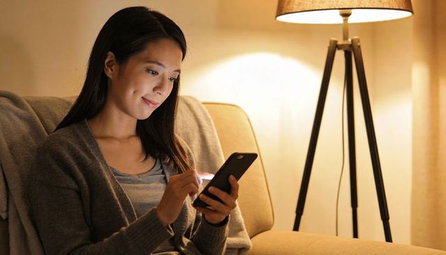 Asian woman relaxing on sofa in evening, scrolling smartphone under warm lamp light