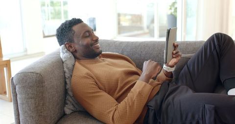 Mid-adult african american man reclining on gray sofa using tablet and smiling