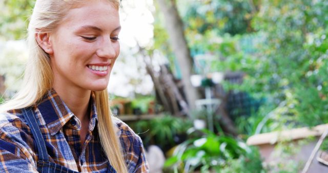 Young blond woman tending to plants in a sunny outdoor yard. She smiles while enjoying gardening tasks. Ideal for articles on outdoor activities, gardening tips, healthy hobbies, joy in nature, or lifestyle blogs.