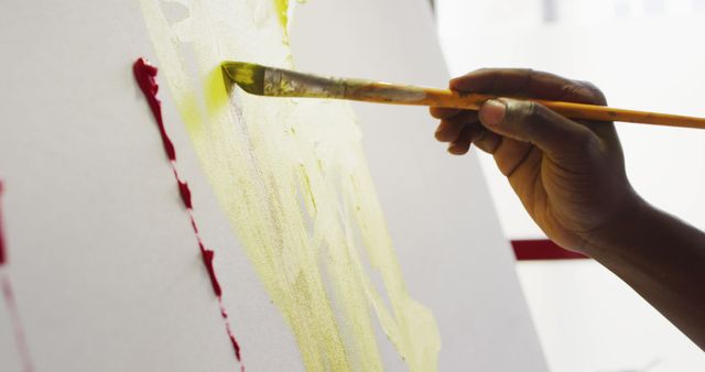 This image features a close-up of an adult hand holding a brush while painting on a canvas with yellow paint. Ideal for use in articles, tutorials, or websites related to art, creativity, and painting techniques.