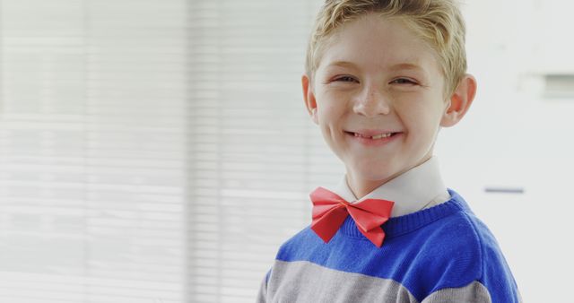 A young Caucasian boy smiles brightly, wearing a vibrant red bow tie and a blue sweater over a collared shirt, with copy space. His cheerful expression and smart attire suggest a special occasion or a formal event.