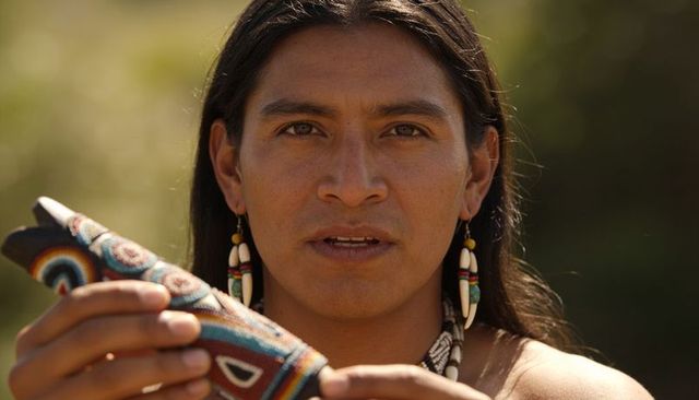 Bare-shouldered man wearing beaded earrings holding handcrafted pouch in sunlight