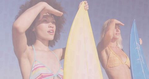 Two women shielding eyes while looking toward ocean holding pastel surfboards in sunny swimwear