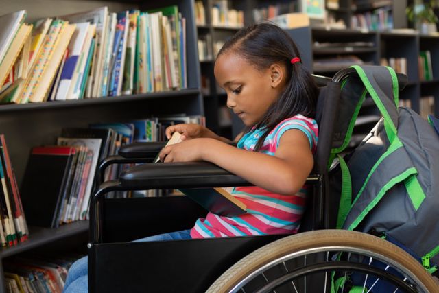 In school, young biracial girl is sitting in a wheelchair, reading a book. She has dark hair tied back, wearing a striped shirt, with a backpack beside her, unaltered
