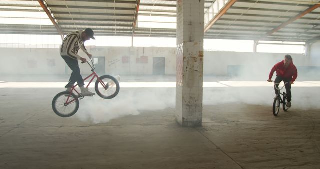 Two young Caucasian men perform bike stunts in a warehouse - Download Free Stock Photos Pikwizard.com