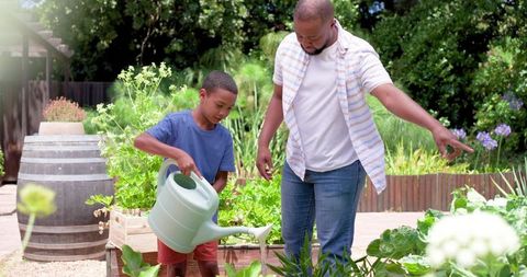 African american father teaching son gardening with watering can in sunny backyard