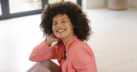 Smiling woman with curly hair relaxing on floor in coral athleisure near glass doors