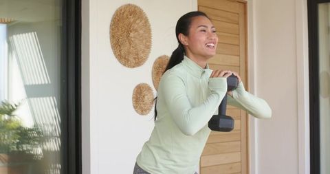 Asian woman exercising with dumbbell on sunlit patio during home strength training session
