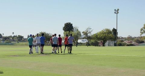 Cricket team walking across suburban sports field carrying bats and pads on sunny day