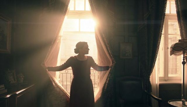 Woman parting heavy drapes revealing sunlit paned window and balcony in vintage parlor