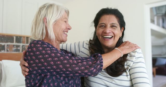Two happy diverse senior women embracing and laughing in sunny bedroom. Friends, retirement, well being, domestic life and senior lifestyle, friendship, unaltered.