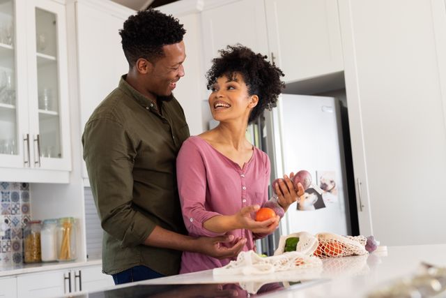 Happy biracial couple embracing, smiling at each other, preparing food in kitchen, copy space. Inclusivity, domestic life, leisure time and togetherness concept.