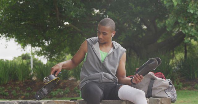 Young man sitting on a stone ledge outdoors putting on sports shoes. Nearby, he has a sports bag and a pair of earbuds in his ears. Lush greenery surrounds him, with trees and bushes in the background. This image can be used for promoting fitness, outdoor activities, sportswear, workout gear, and healthy lifestyle campaigns.