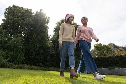 Two friends wearing santa hats walking across park lawn, knit sweaters, holiday stroll