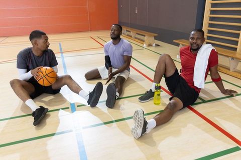 Three male teammates resting on gym floor, chatting and rehydrating after basketball pickup