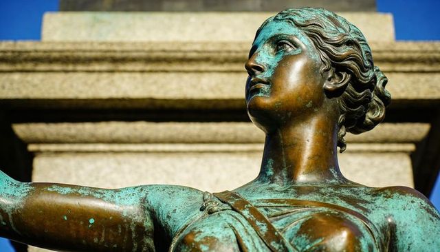 Sunlit bronze female statue with verdigris patina on pedestal, classical greco-roman detail