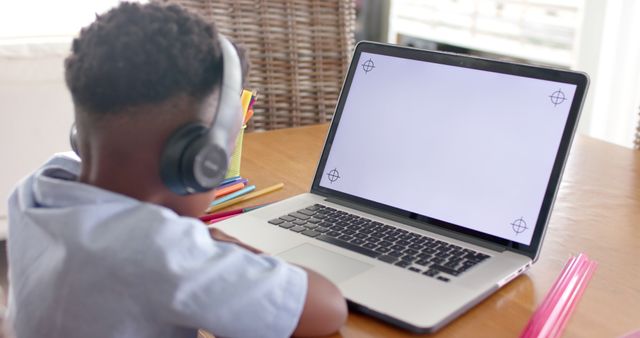 Boy sitting at table, wearing headphones, using laptop at home. Ideal for articles on online education, technology in learning, and resources for remote schooling. Can be used in educational websites, blogs, and social media promoting remote learning and children's use of technology for school.