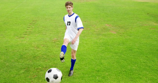 The image depicts a young male soccer player in white and blue sportswear kicking a soccer ball on a green field. This image can be used for sports articles, fitness magazines, promotional materials for soccer clubs, and athletic gear advertisement.