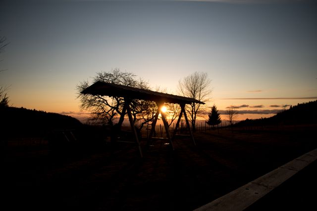 Capturing the serene moment of sunset behind a wooden shelter and bare trees in a rural landscape. The silhouette effect creates a tranquil atmosphere, making it perfect for themes related to peace, nature, and tranquility. Use this image for websites, blogs, or promotional materials focusing on relaxation, rustic travels, or outdoor lifestyle.