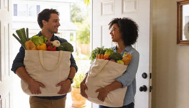 Smiling couple carrying reusable grocery bags brimming with fresh produce at home doorway
