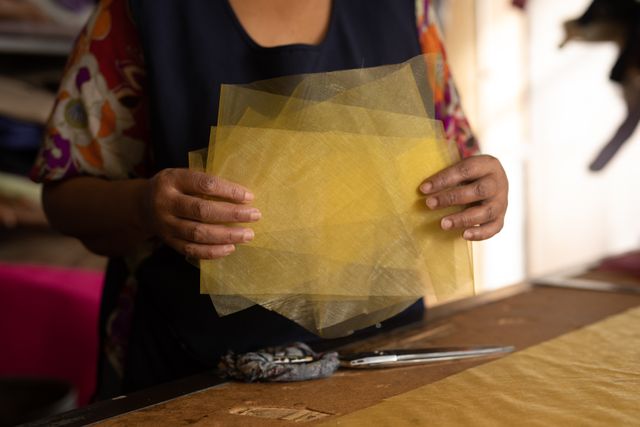 Biracial woman holding yellow tissue fabric in a hat factory, showcasing the crafting process. Ideal for use in articles about traditional crafts, small businesses, textile industry, and handmade products. Can be used in blogs, websites, and promotional materials highlighting artisanal work and manufacturing processes.