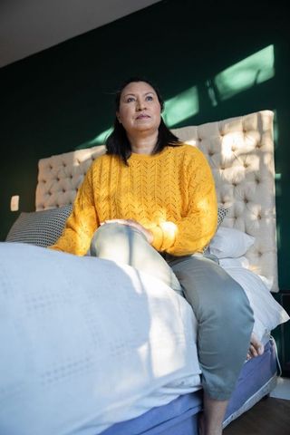 Middle-aged woman sitting on bed edge wearing mustard sweater gazing into morning sunlight