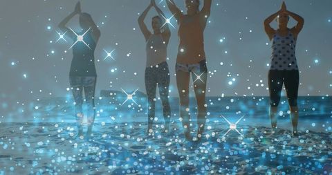Four women practicing sunrise beach yoga in prayer pose with blue bokeh sparkle overlay