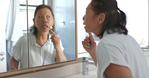 Senior asian woman using daily facial cleansing brush while checking skin in vanity mirror